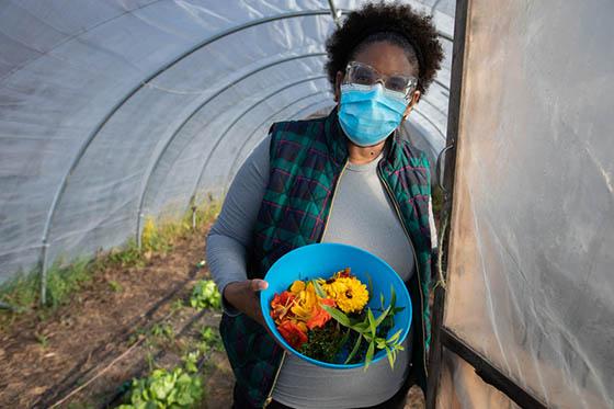 Photo of a masked Chatham University student holding a blue bowl of harvested produce, at the entrance to a greenhouse on Eden Hall Campus