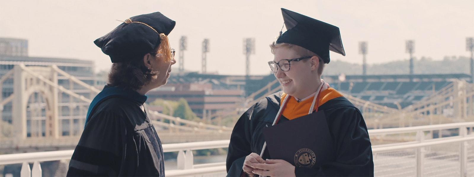Photo of a Chatham University graduate laughing with a professor in front of a Pittsburgh skyline with yellow bridges. 