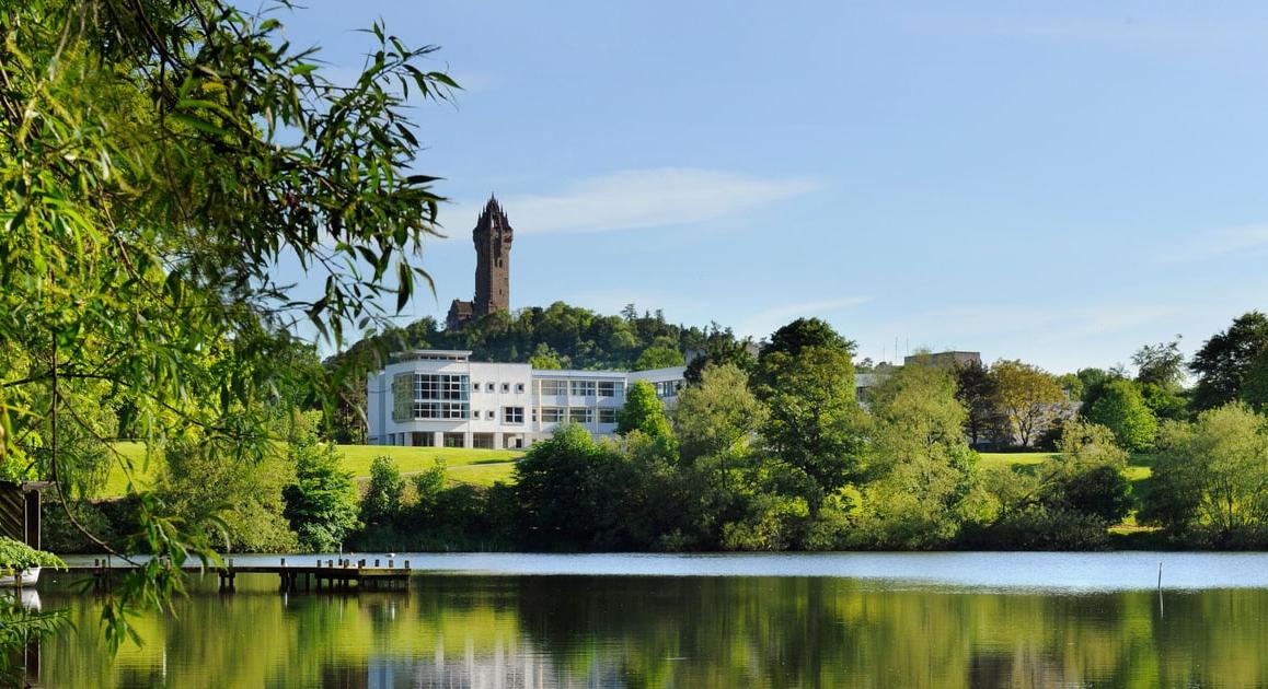 Photo of green forests and a calm lake, with a white building and a tall tower behind it: The University of Stirling in Stirling, Scotland.