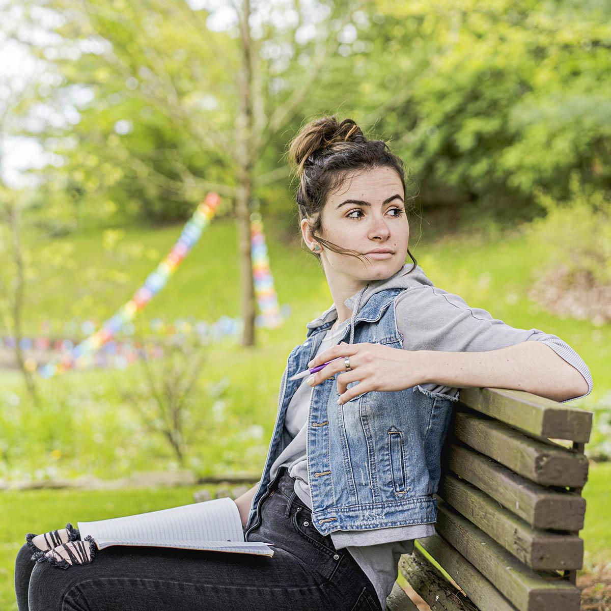 Photo of a young white woman with hair tied up, wearing a jean jacket and seated on a bench outside looking behind her into the distance